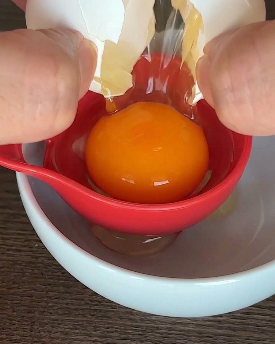A raw egg being separated in a red tool over a white bowl, with the orange yolk held in the center.