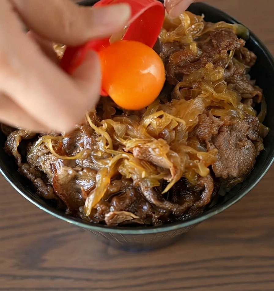 A bright orange egg yolk being dropped from a red separator onto the center of a finished beef bowl.