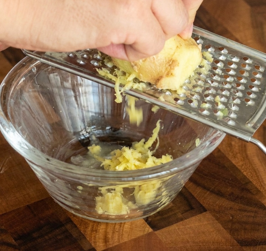 A hand rubbing a piece of fresh ginger against a metal grater, with the paste falling into a small clear glass bowl.