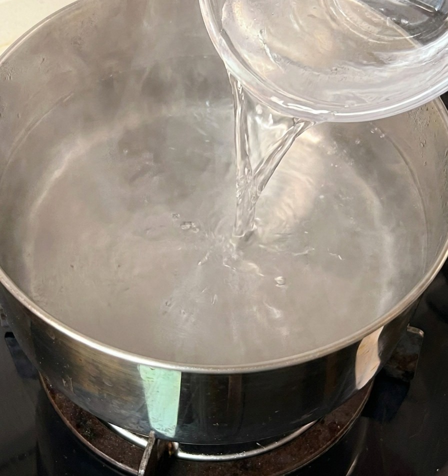 Pouring a small glass bowl of cold water into a stainless steel pot of hot water.