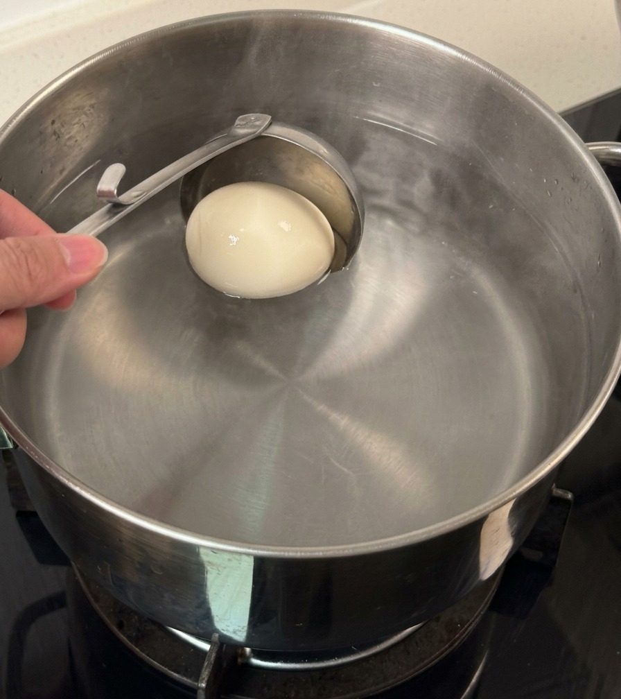A metal ladle lowering a single whole white egg into a pot of water.