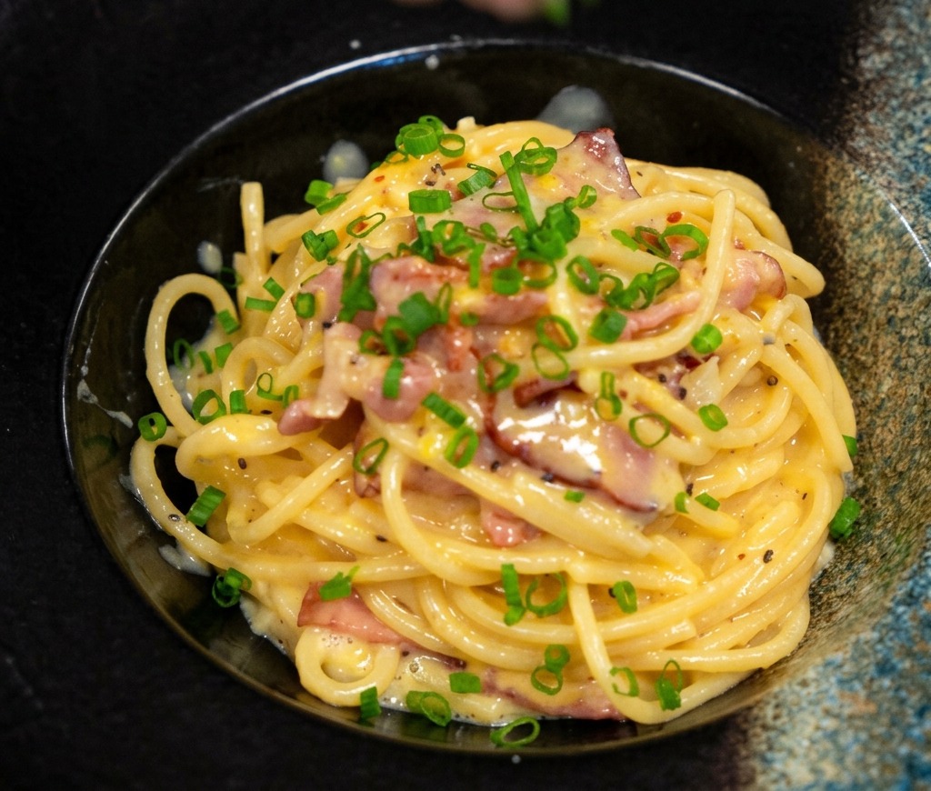 Fingers sprinkling bright green chopped scallions over a beautifully plated bowl of creamy bacon pasta.