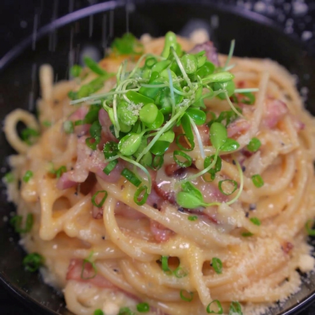 Fresh cheese snowing down onto a finished bowl of creamy bacon pasta garnished with green onions and microgreens.