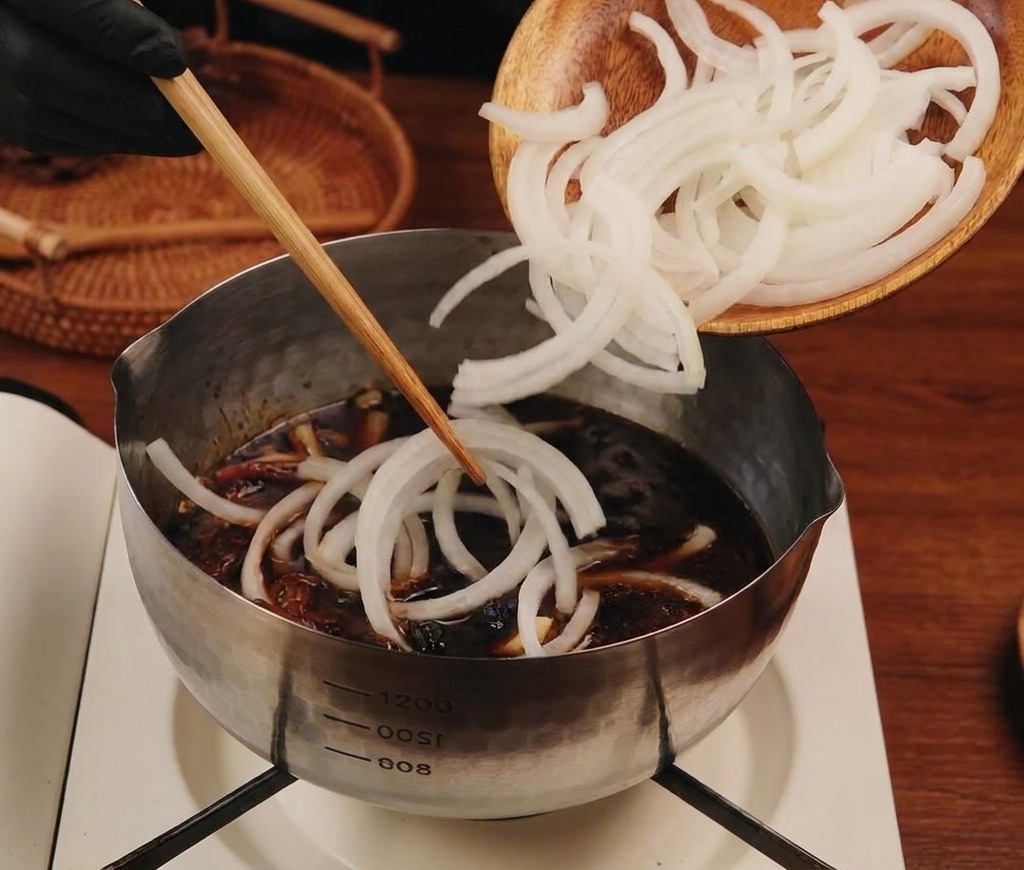 Sliced white onions being dropped from a wooden plate into a stainless steel pot filled with dark savory liquid.