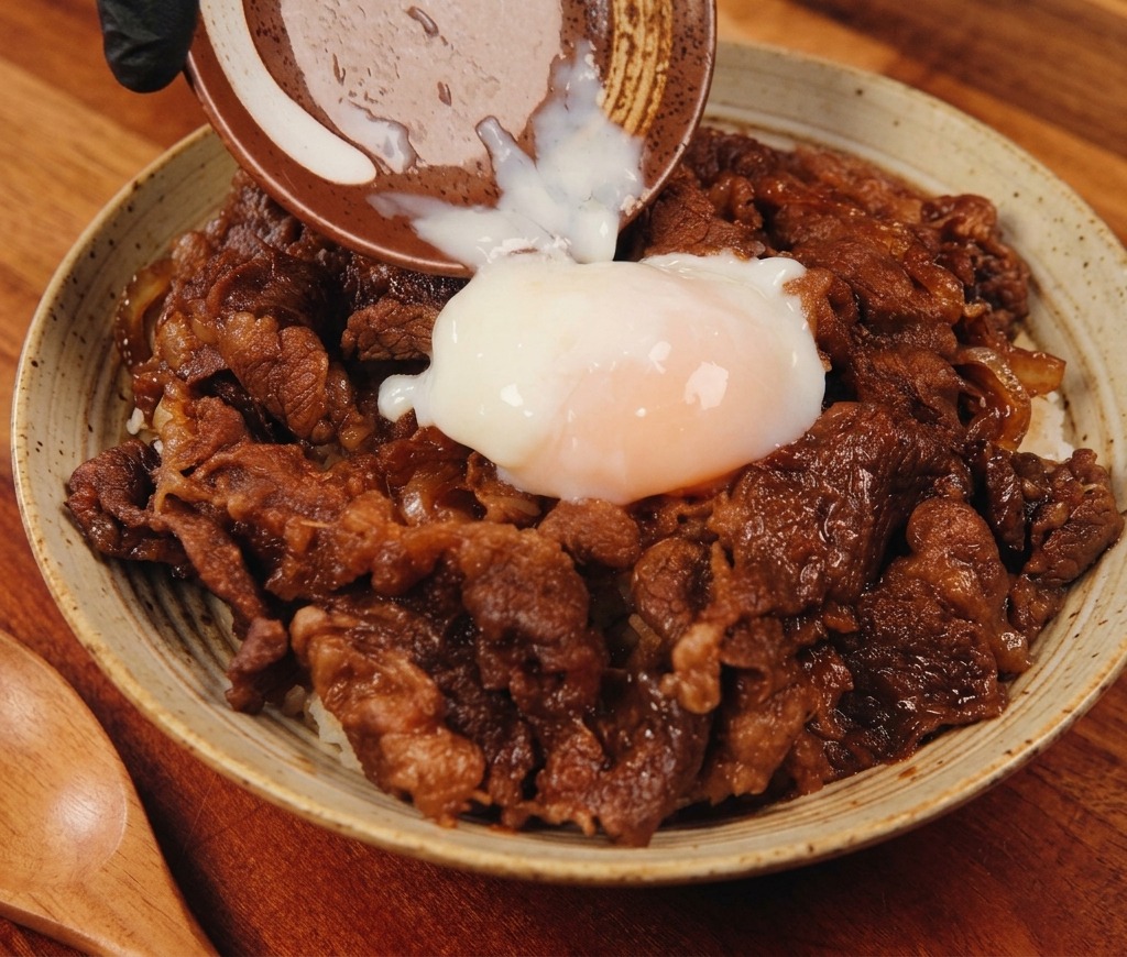 A soft-boiled onsen egg being poured from a small ceramic dish onto a steaming bowl of savory beef and rice.