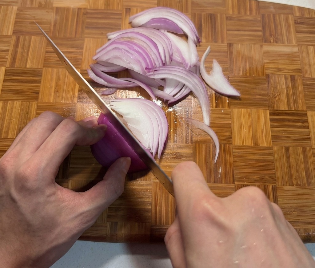 A pair of hands slicing a red onion into thin wedges on a bamboo cutting board with a sharp chefs knife.