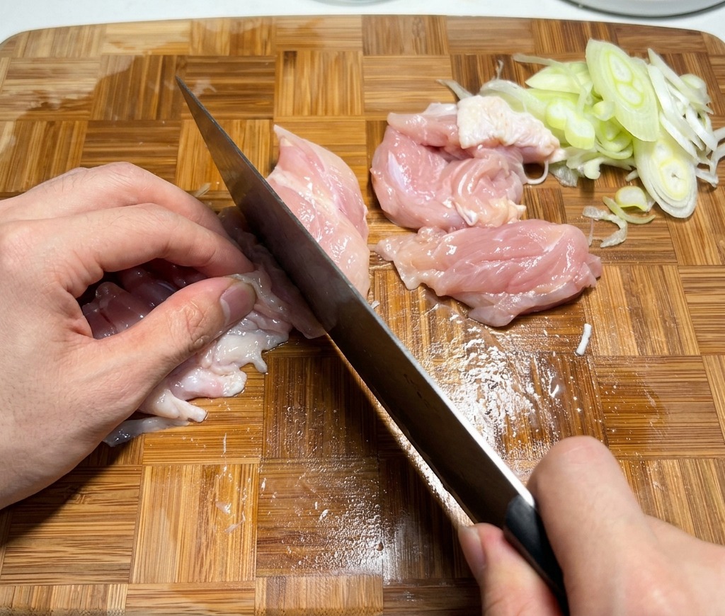 Slicing raw chicken thighs into small pieces on a wooden cutting board.