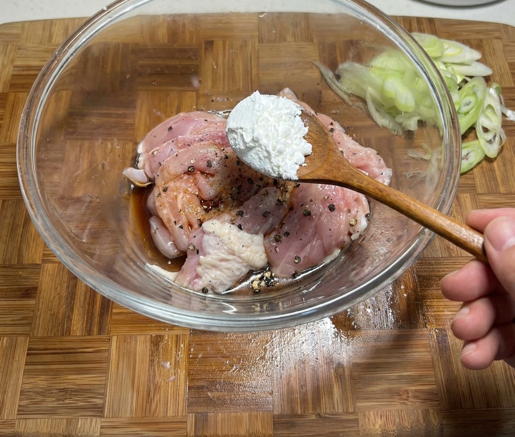 Adding a spoonful of cornstarch to a bowl of raw chicken marinated in soy sauce and spices.