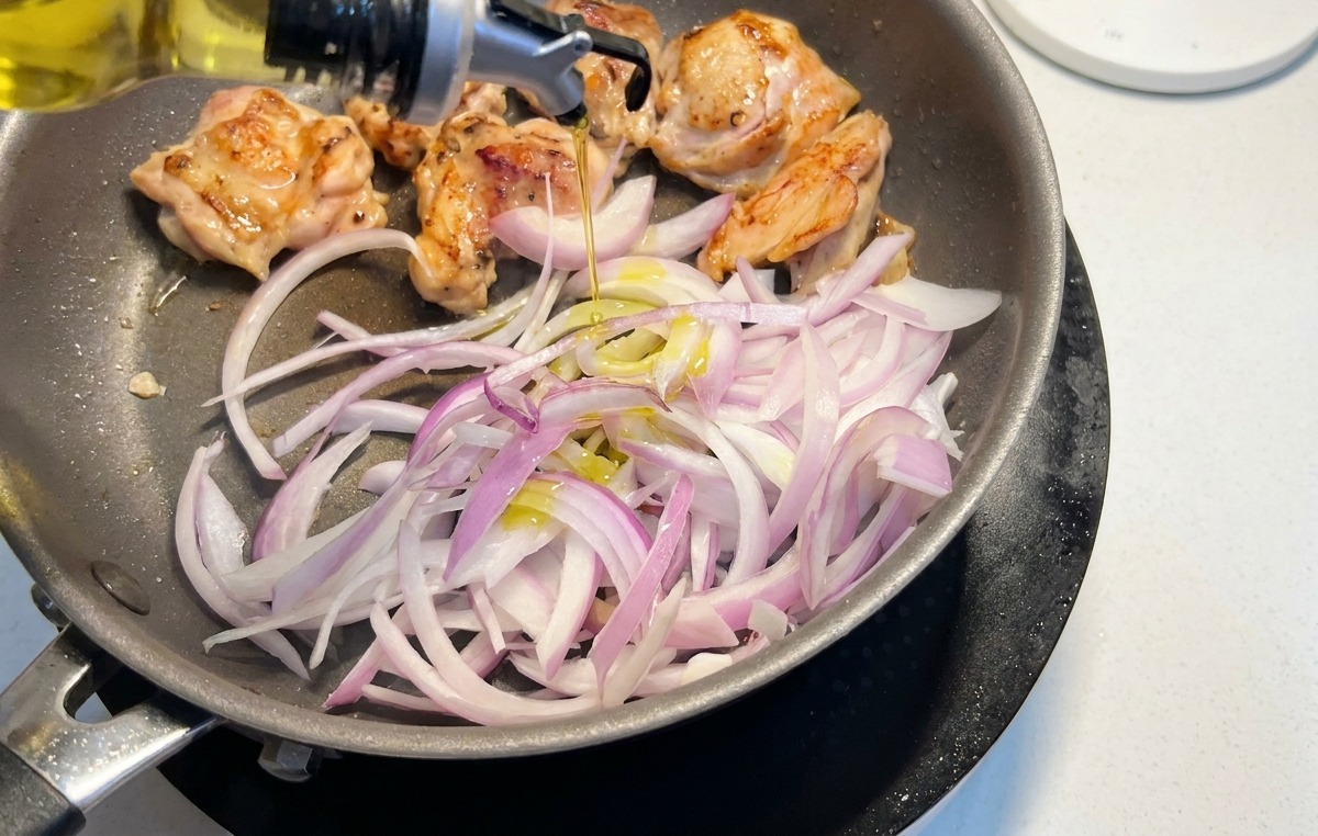 Sliced red onions being added to a pan with browning chicken thighs while oil is drizzled over them.