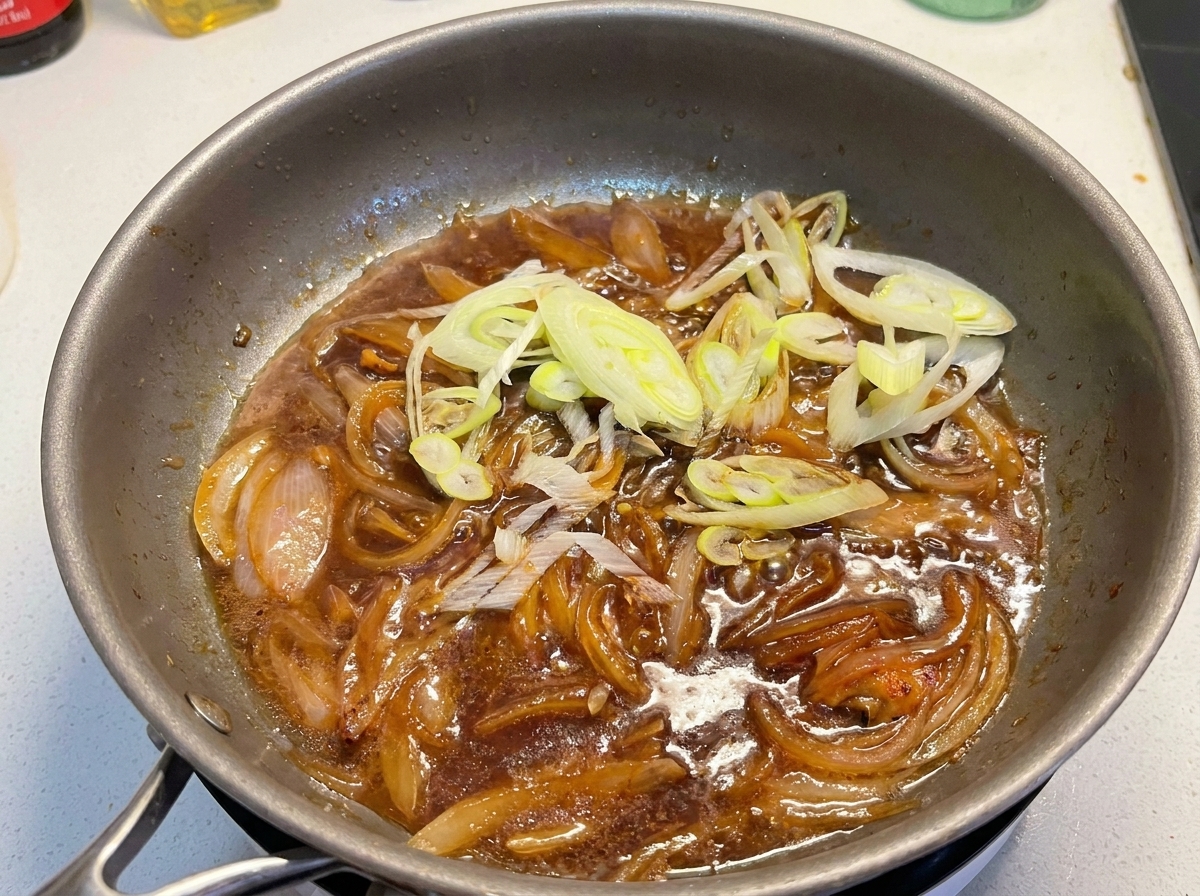 Sliced scallions being added to a pan containing a dark simmering sauce and cooked onions.