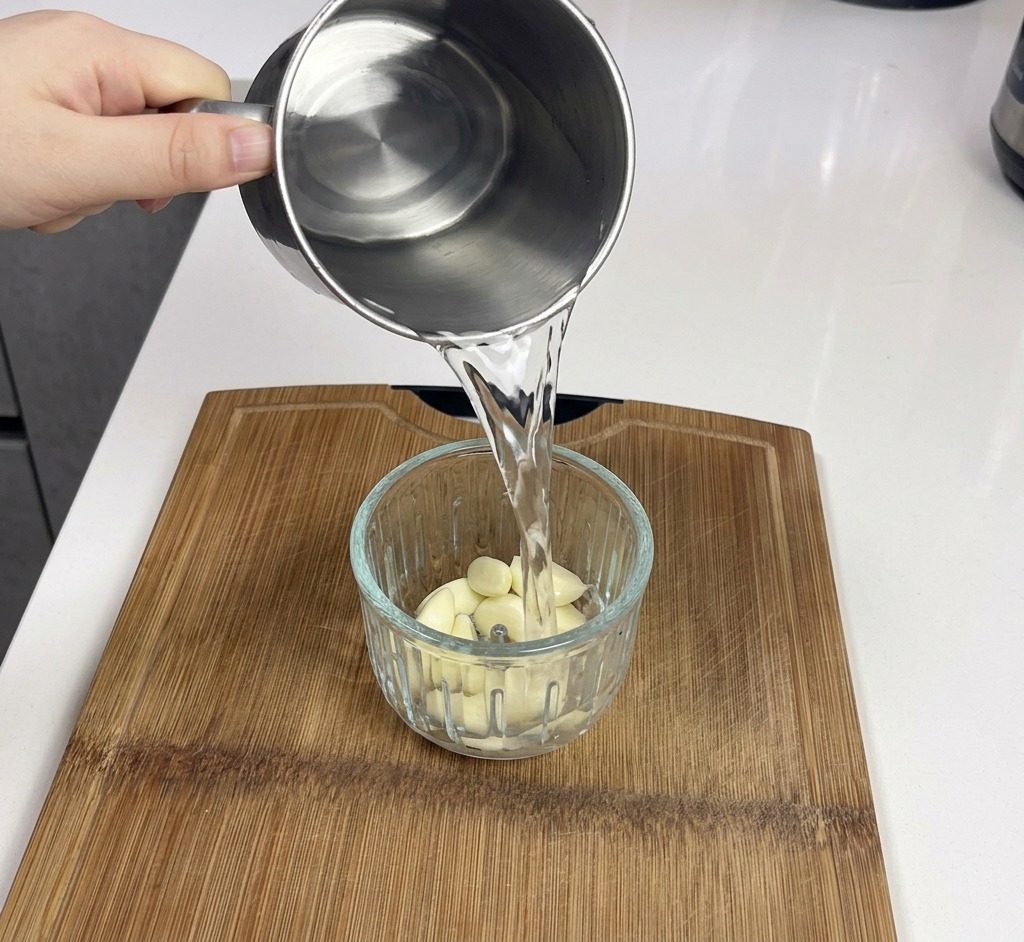 A metal mug pouring water into a clear glass blender jar filled with fresh white garlic cloves.