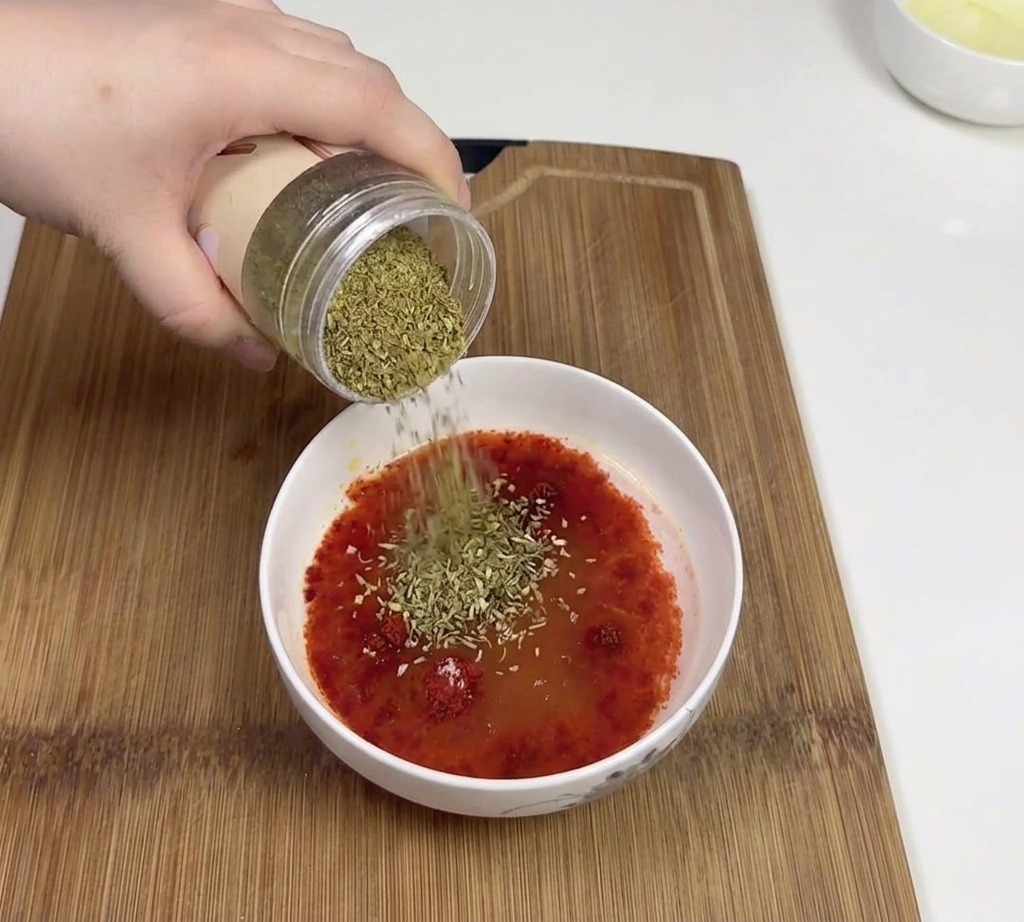 Hand pouring dried green cumin powder from a jar into a bowl containing a red chili sauce base.