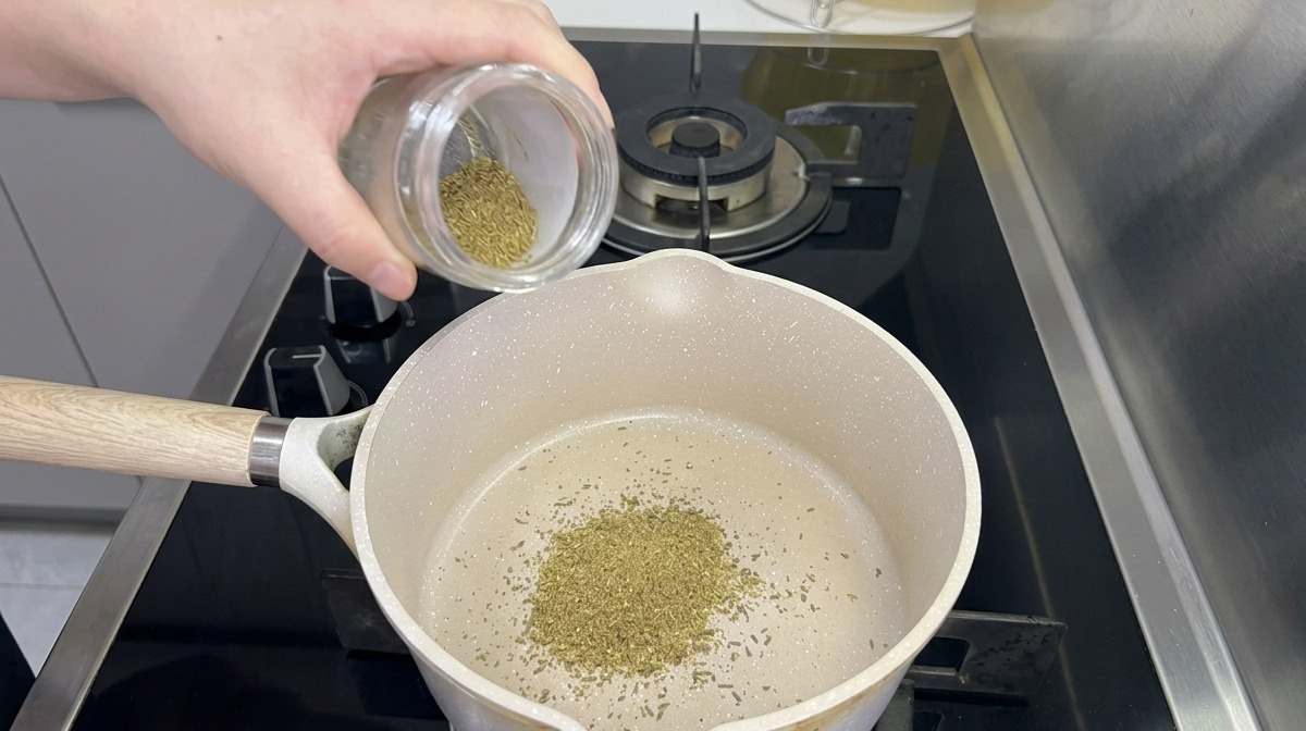 Cumin powder being poured from a glass jar into a white saucepan resting on a black gas stove.