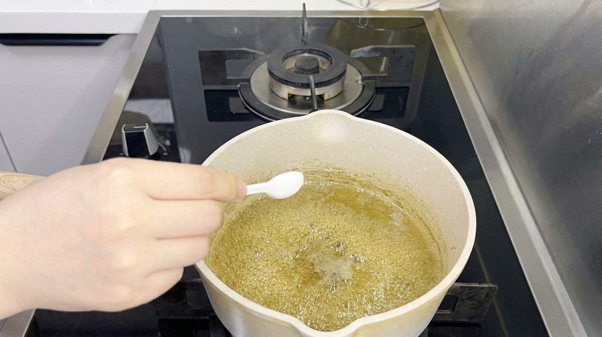 A person using a small white spoon to add seasoning to a boiling, greenish-tinted liquid in a saucepan.