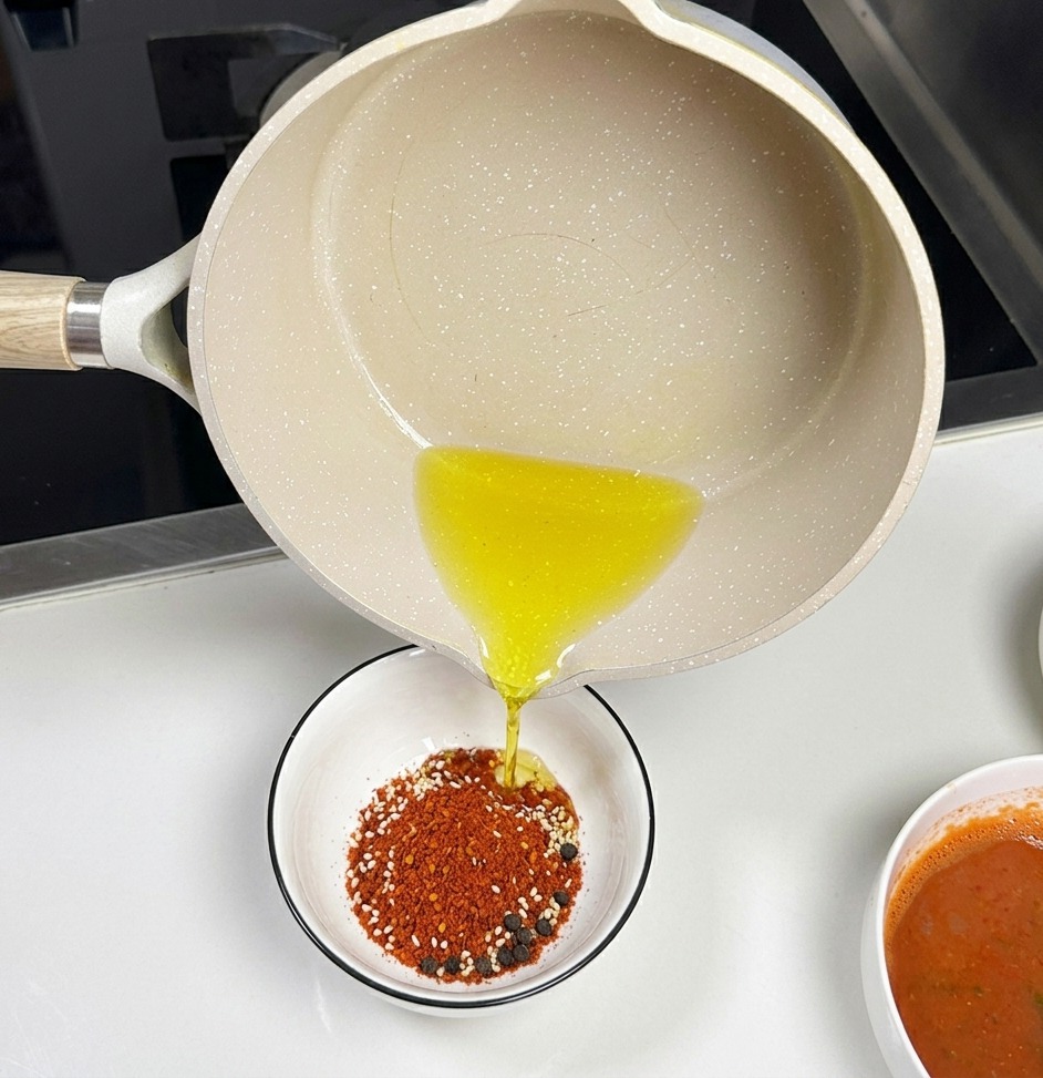 A light-colored ceramic saucepan pouring clear hot oil into a small white bowl filled with red chili powder and white sesame seeds.