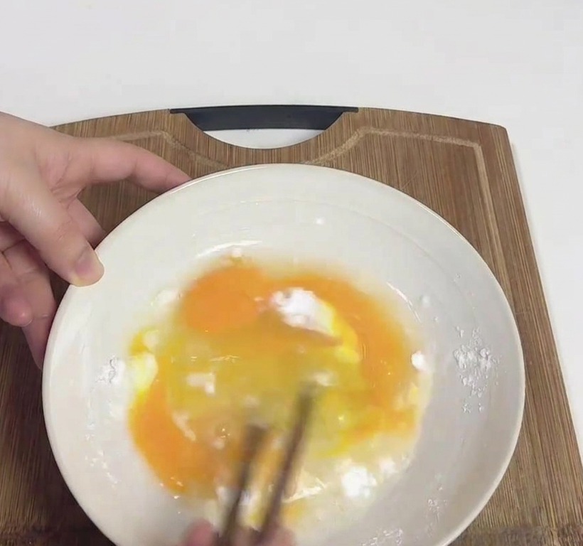 Hands whisking yellow eggs and white potato starch in a white bowl using a pair of wooden chopsticks.
