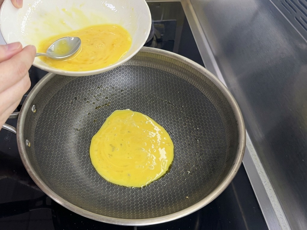 Thin yellow egg batter being poured from a bowl into a large hexagonal-patterned frying pan.