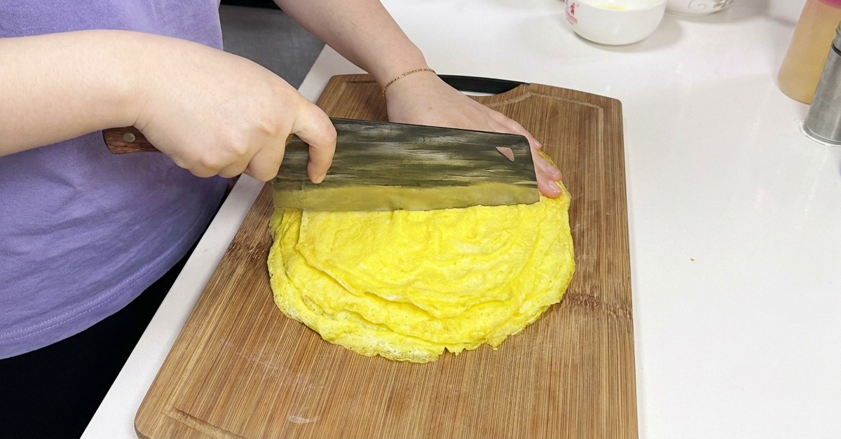 A person using a large Chinese cleaver to cut through a stack of thin, cooked yellow egg pancakes on a wooden board.