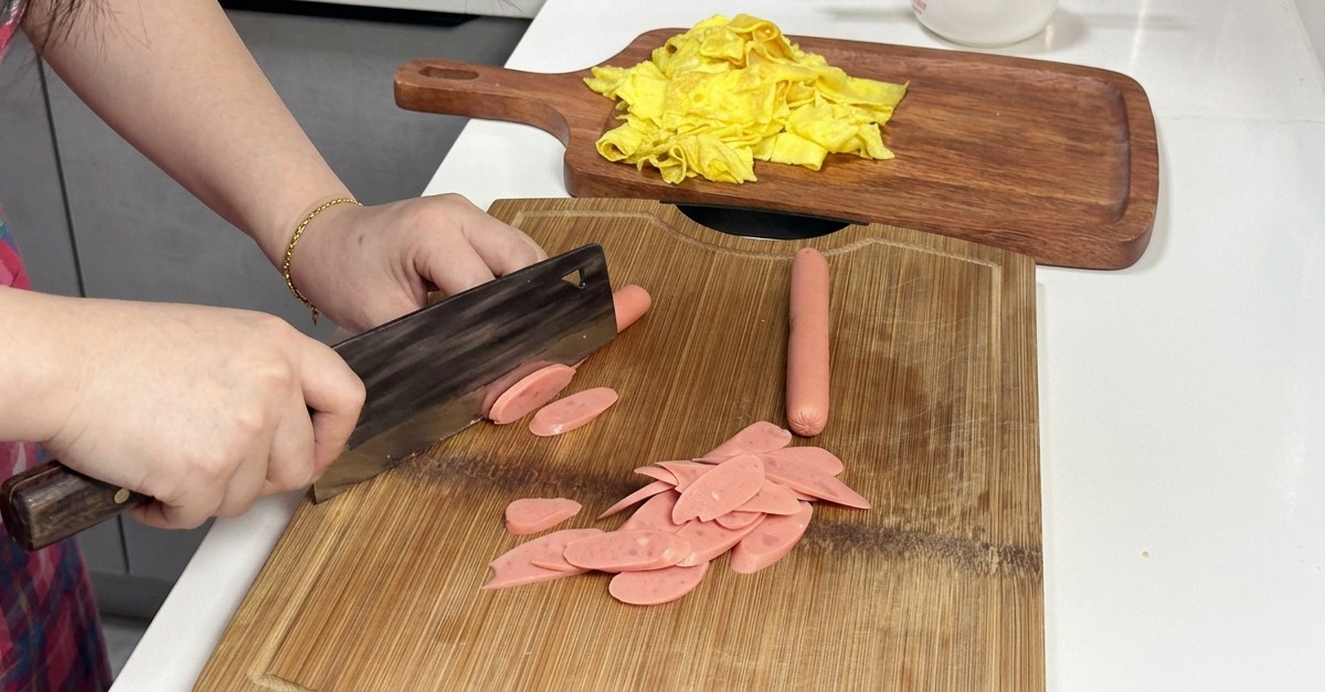 A person slicing pink sausages diagonally on a wooden cutting board with a cleaver.