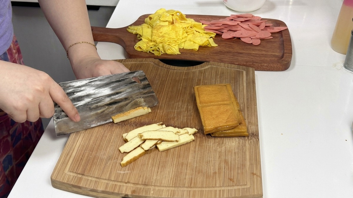 A metal cleaver slicing through a stack of golden-brown smoked bean curd blocks on a wooden cutting board, with prepared egg and ham strips in the background.