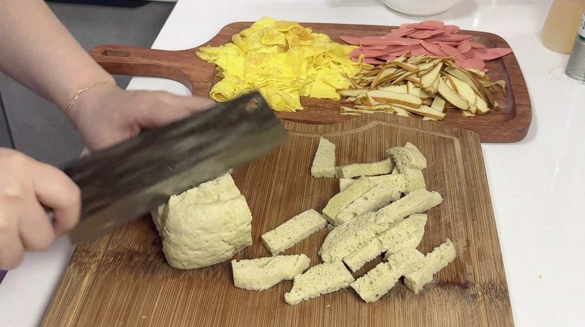 A large block of pale, spongy steamed gluten being sliced into rectangular portions on a wooden board using a cleaver.