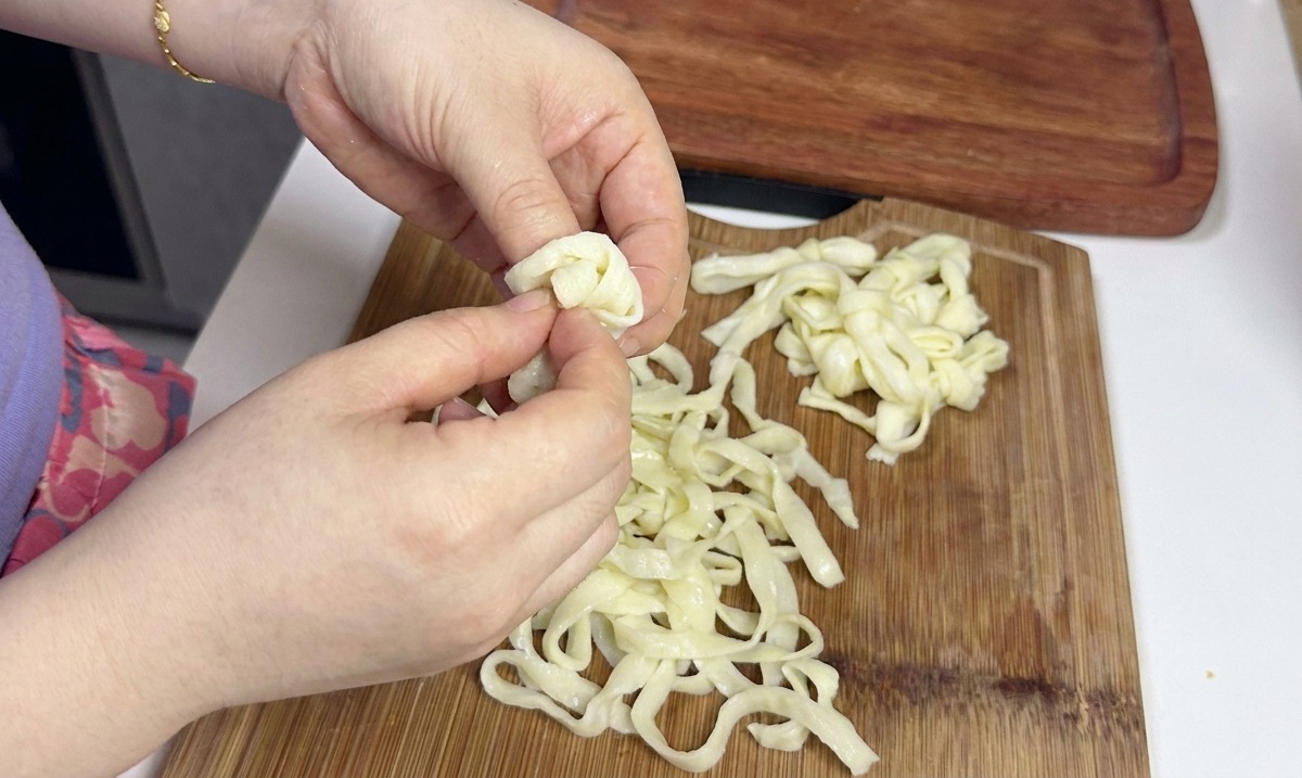 Hands carefully separating and loosening a pile of wide, flat white cold noodles on a wooden cutting board.