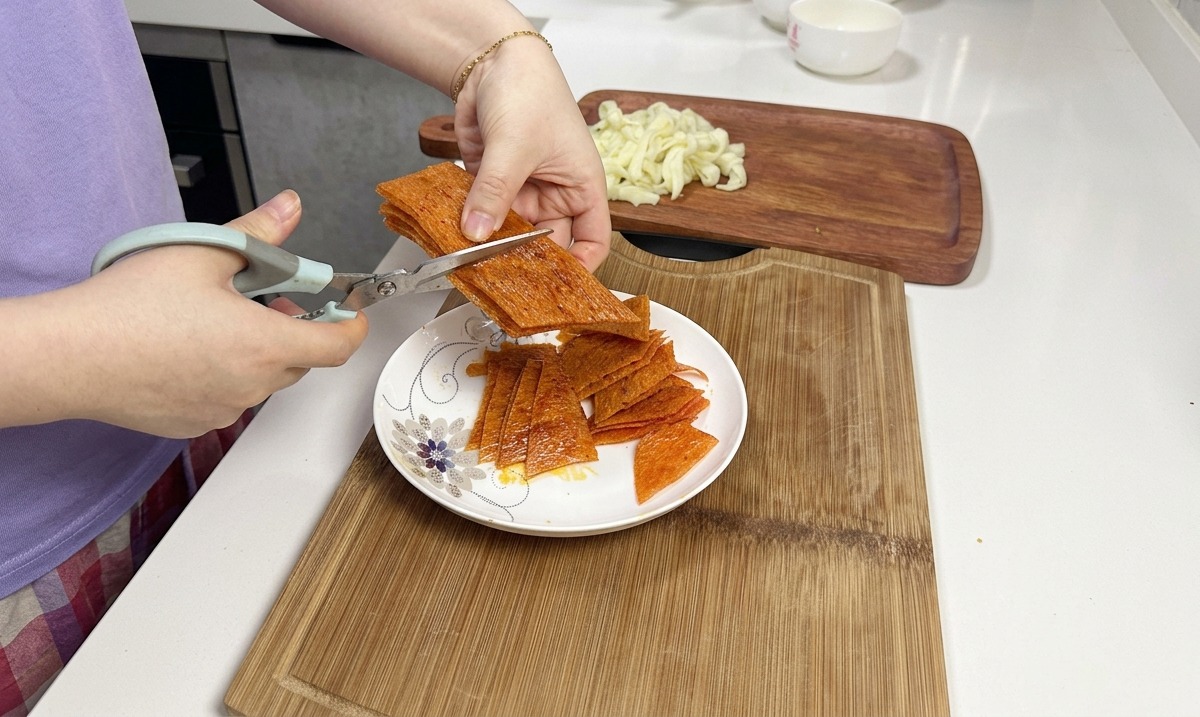 Kitchen scissors cutting through a stack of orange spicy latiao strips over a white ceramic plate.