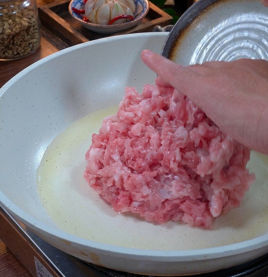 Fresh pink ground pork being placed into a white ceramic frying pan with a light coating of oil.