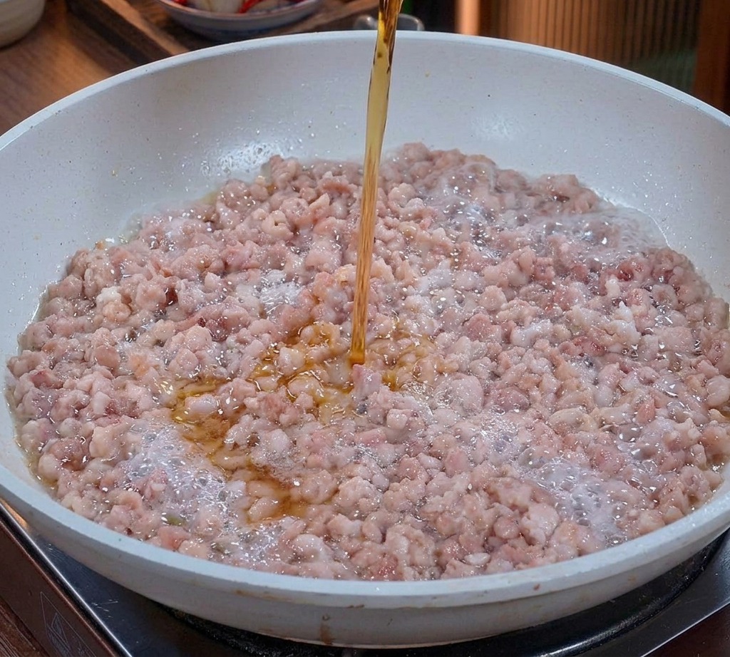 A clear amber liquid being poured from a glass onto stir-frying minced pork in a pan.