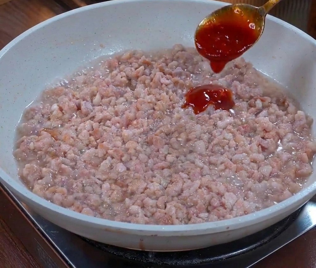 A metal spoon pouring a thick, dark brown sauce onto a bed of stir-fried ground meat in a pan.