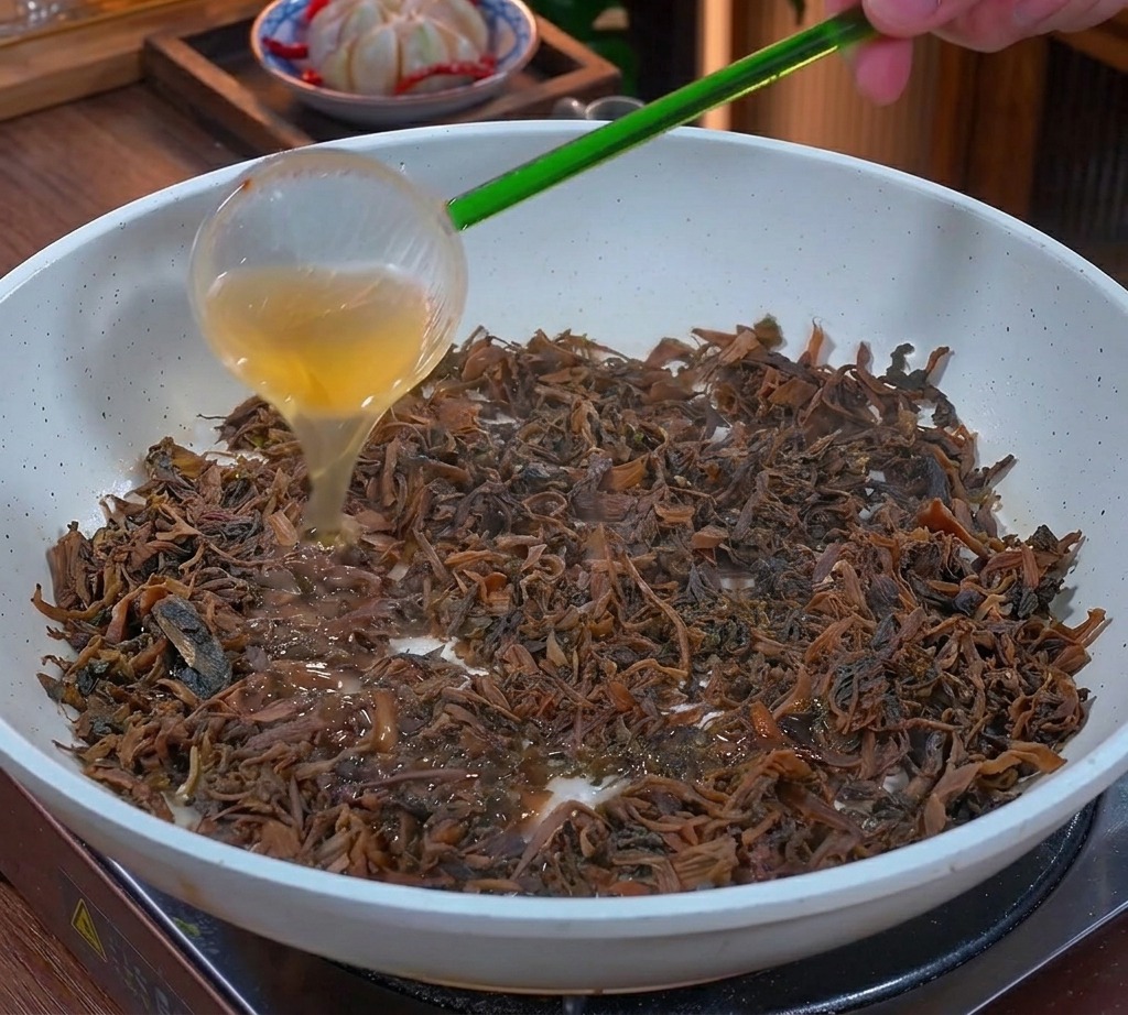 Golden liquid fat being poured from a green ladle over sautéed chopped mustard greens in a white pan.