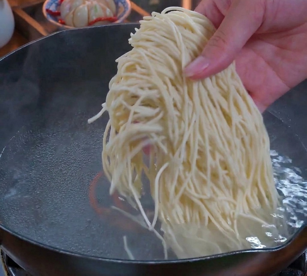 A hand dropping a thick bundle of thin, fresh noodles into a pot of boiling water.