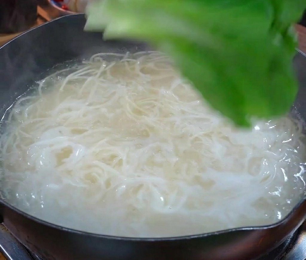 Fresh green leafy vegetables being added to a pot of boiling water containing white noodles.