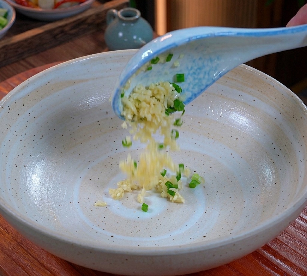 Minced garlic and chopped scallions falling from a blue and white spoon into the bottom of a wide ceramic bowl.