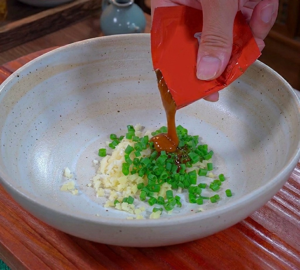 A hand squeezing thick brown Dandan sauce from a red packet into a ceramic bowl over chopped garlic and scallions