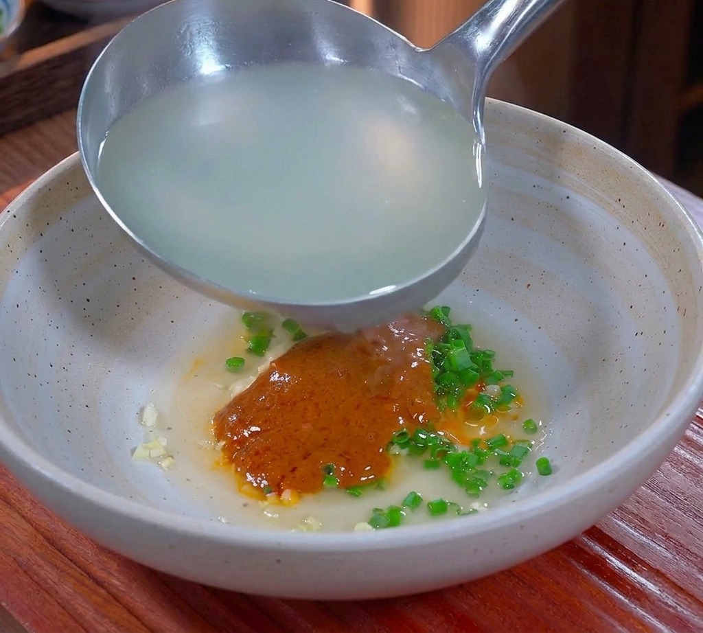 Cloudy hot noodle water being ladled into a bowl containing spicy seasonings and pork fat.
