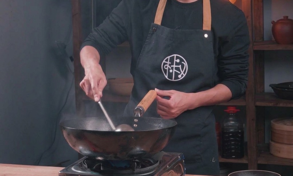 A chef stir-frying minced pork in a traditional black wok adding liquid seasoning to the mixture as it cooks.