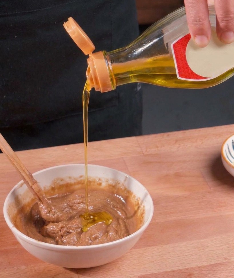Clear golden oil being poured from a bottle into a white bowl containing thick brown sesame sauce while being stirred with a chopstick.