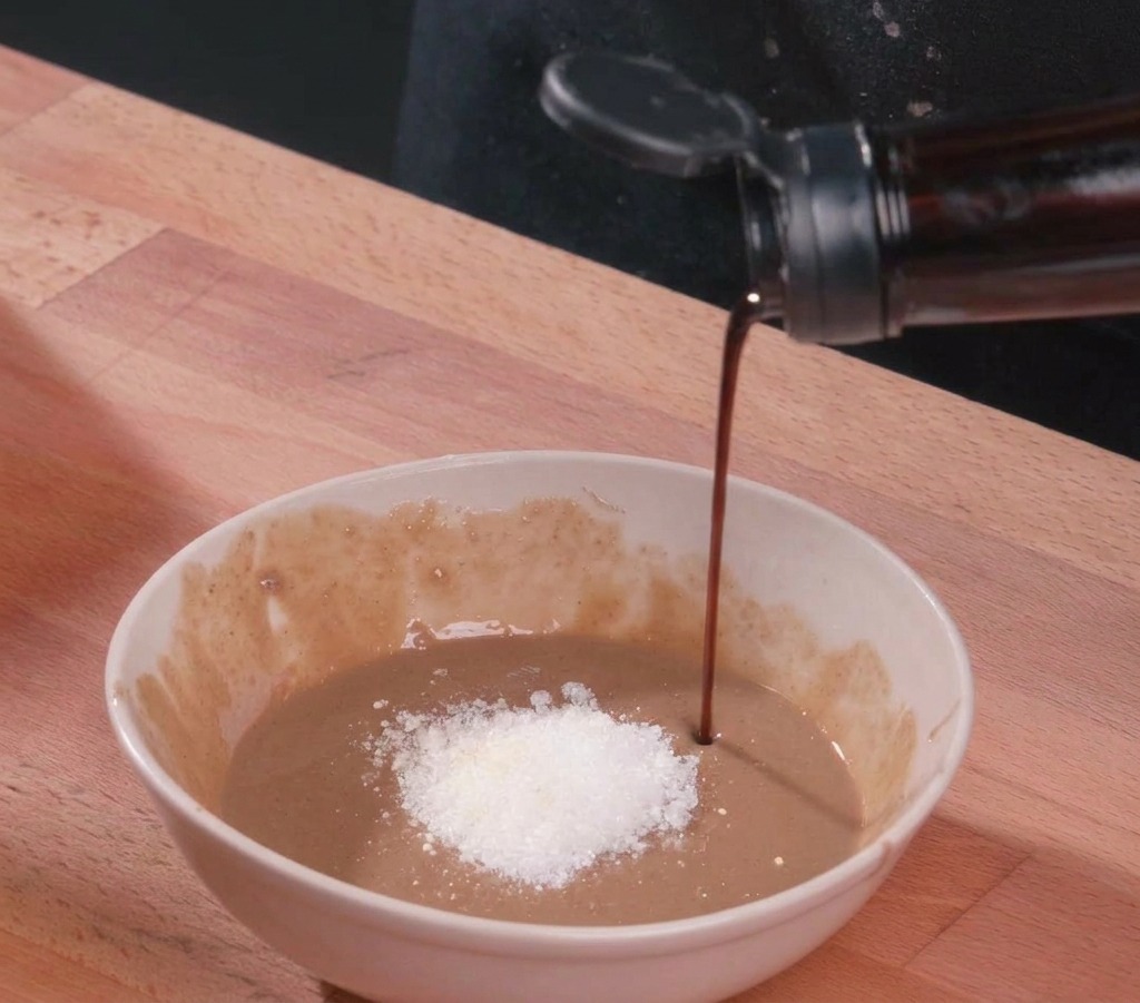 A stream of dark soy sauce being poured into a white ceramic bowl containing thick sesame paste, a mound of white sugar, and vinegar.