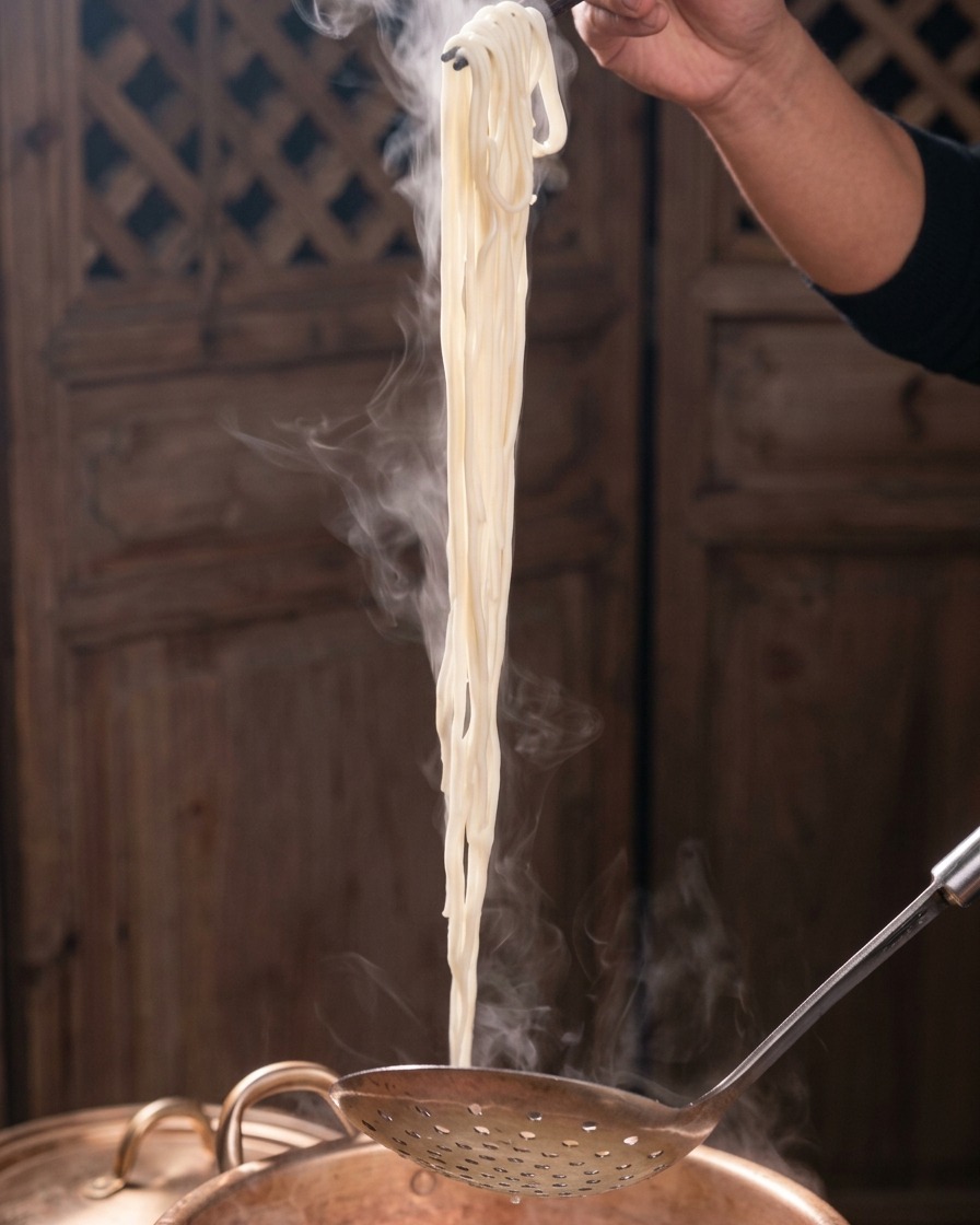 A bundle of steaming white noodles being lifted vertically out of a copper pot using dark chopsticks and a perforated metal ladle.