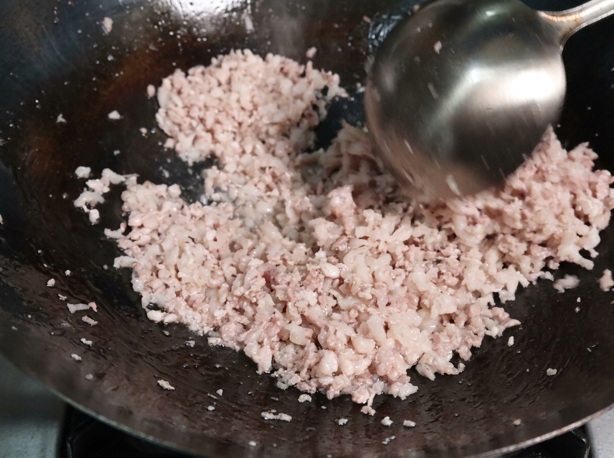 Minced pork being stir-fried with a metal ladle in a hot wok, showing the meat beginning to cook and separate into small grains.