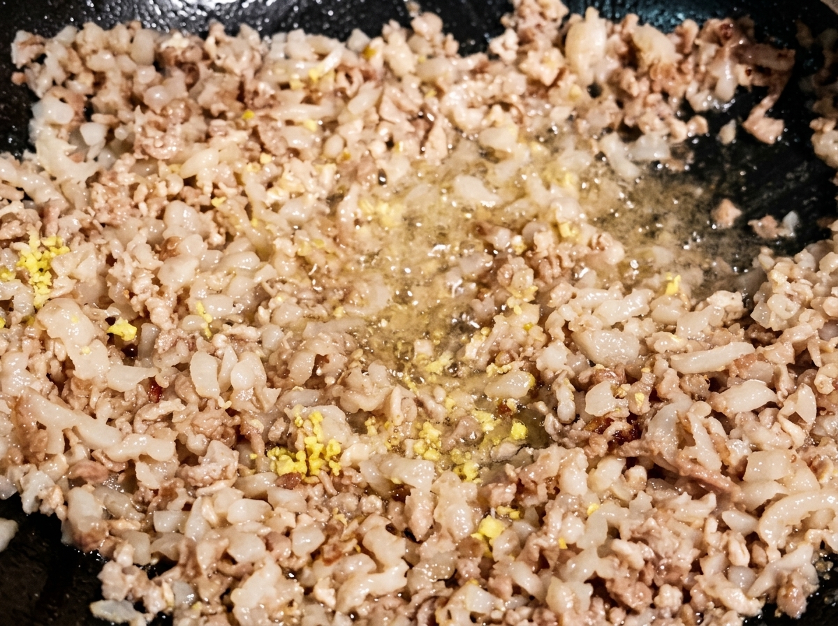 Minced ginger and garlic pieces being stir-fried into browned minced meat in a dark wok.