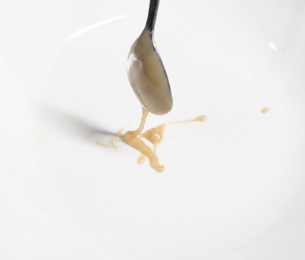A metal spoon pouring thick, light brown sesame paste into a white bowl, marking the first step of creating the noodle sauce base.