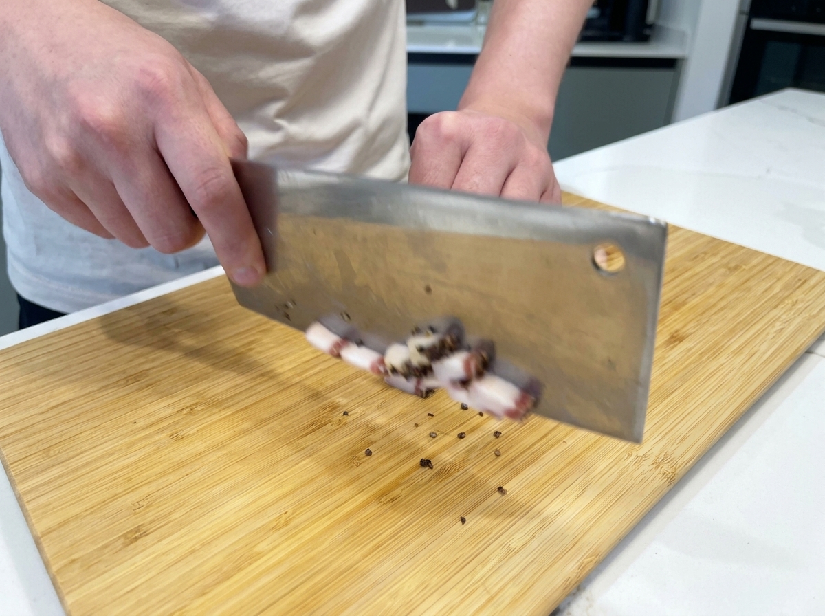 A chef using a large cleaver to cut fatty strips of cured guanciale into small cubes on a bamboo cutting board.