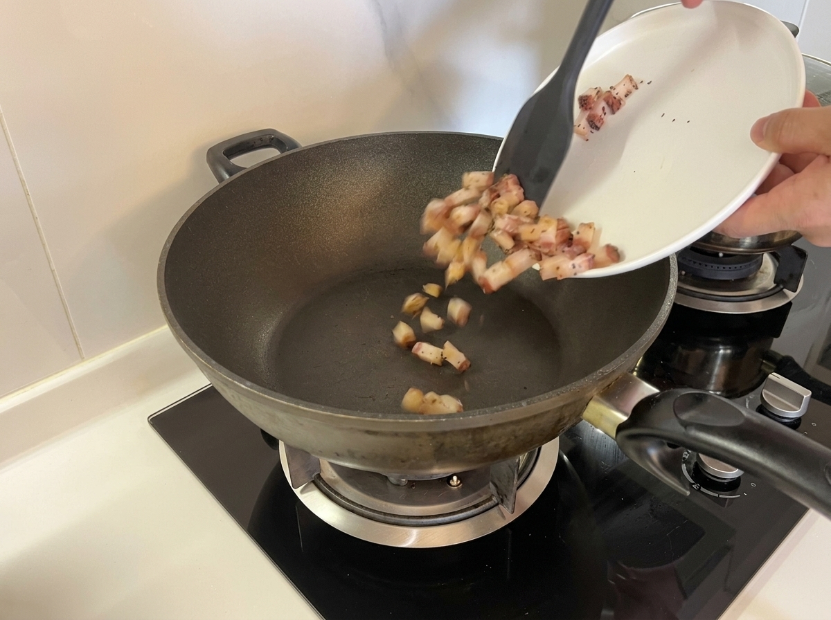 Diced raw guanciale being poured from a white plate into a dry, unheated wok using a spatula.
