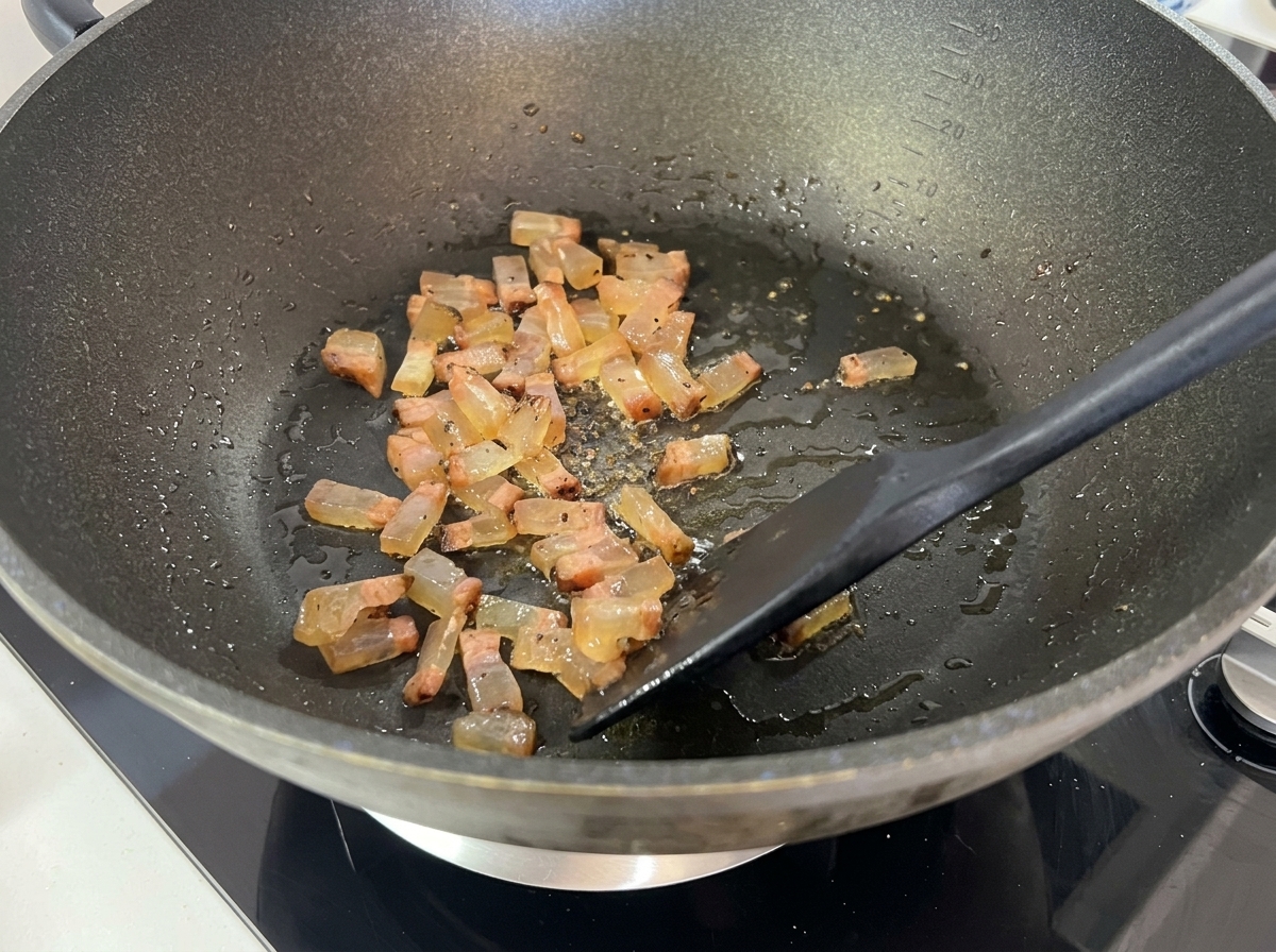 Diced guanciale browning and turning translucent in a wok, actively releasing liquid fat.