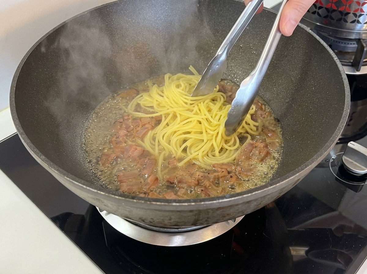 Metal tongs transferring strands of cooked spaghetti into a wok filled with crispy guanciale and a shallow pool of hot liquid.