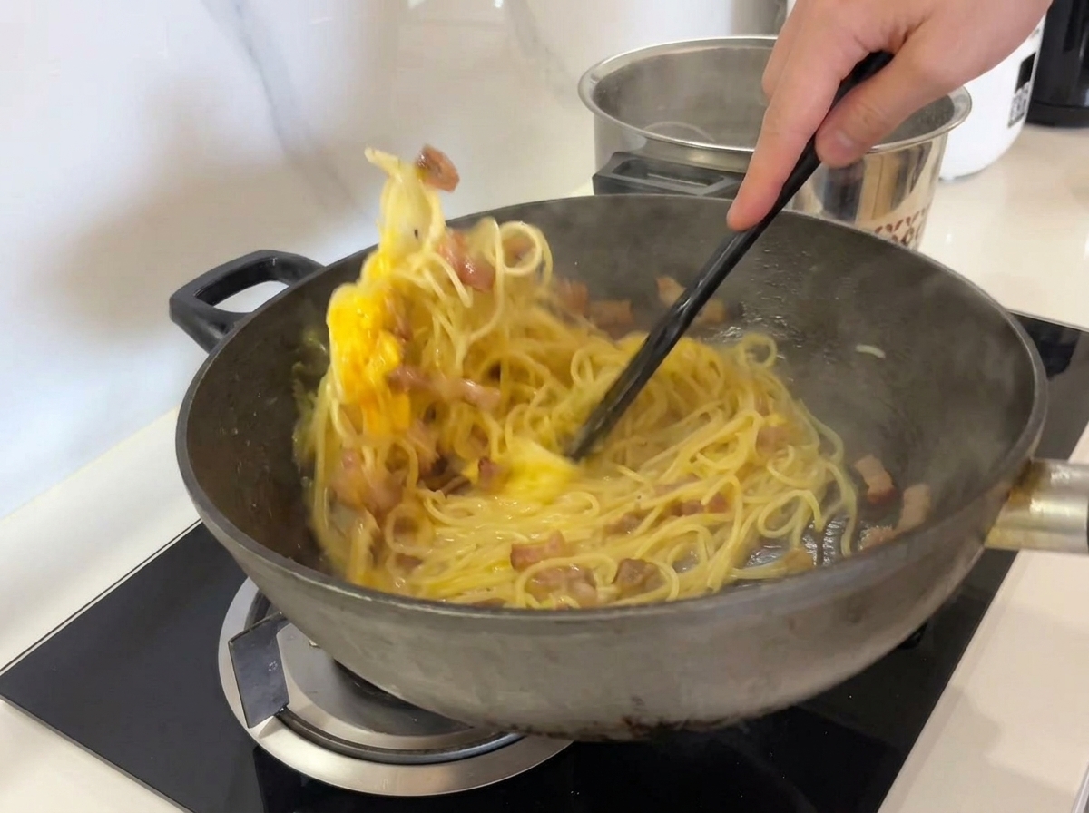 Vigorously stirring spaghetti, guanciale, and the yellow egg mixture in a wok with a black spatula.