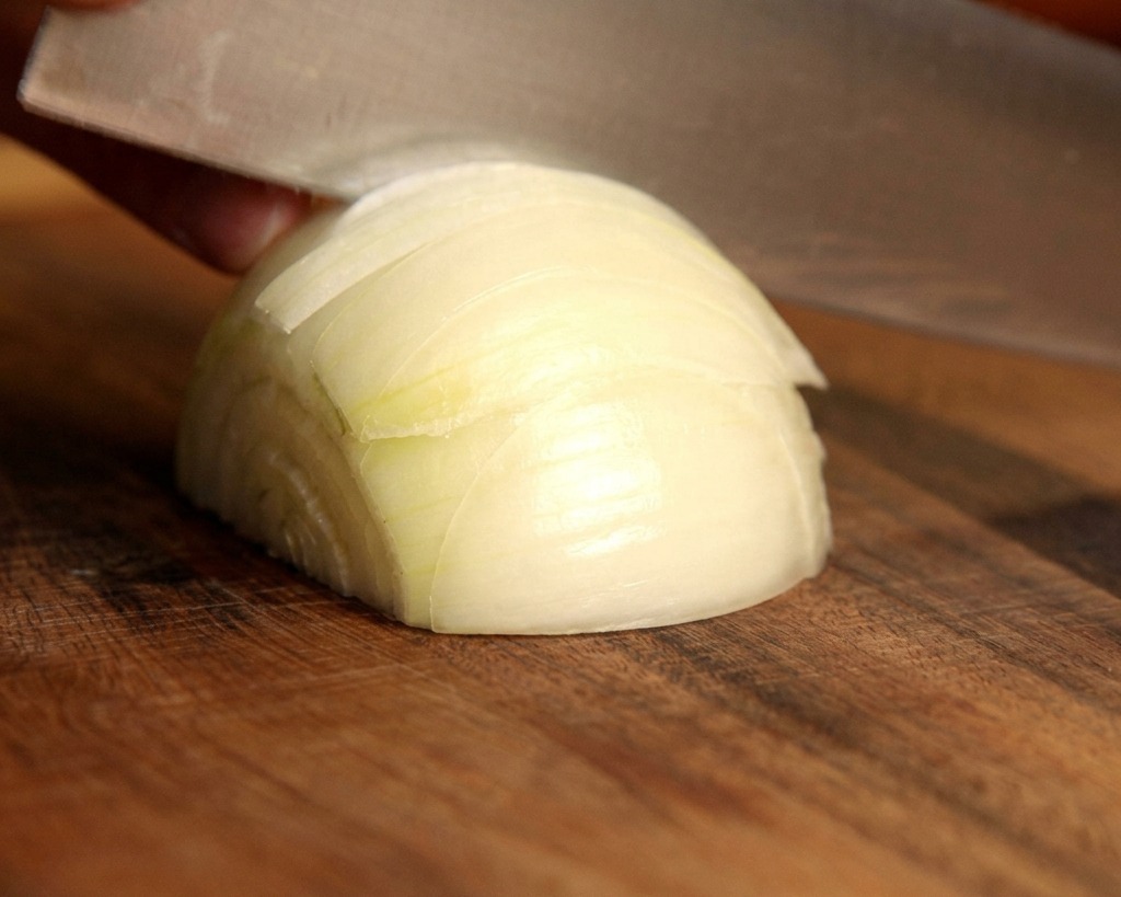 A sharp knife slicing through a fresh white onion to create thin wedges on a wooden cutting board.