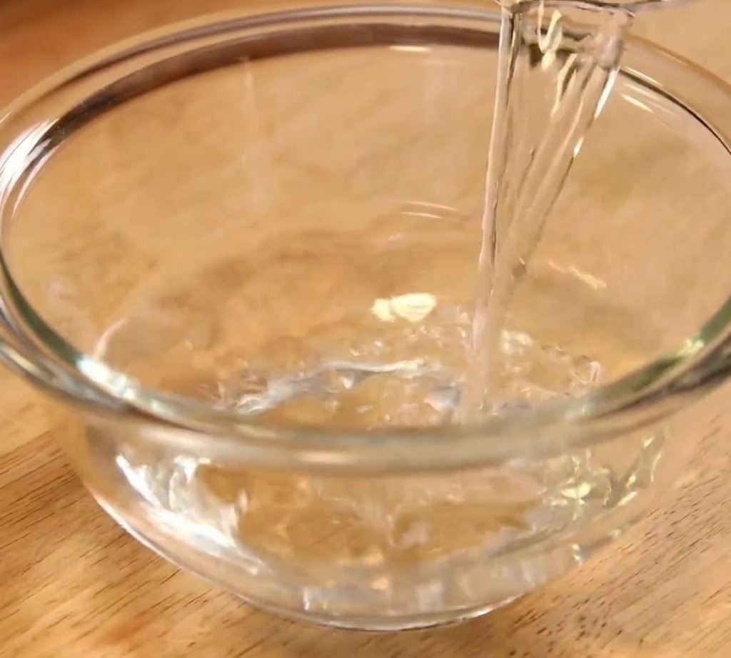 Clear water being poured into a transparent glass mixing bowl as the first step of the sauce preparation.